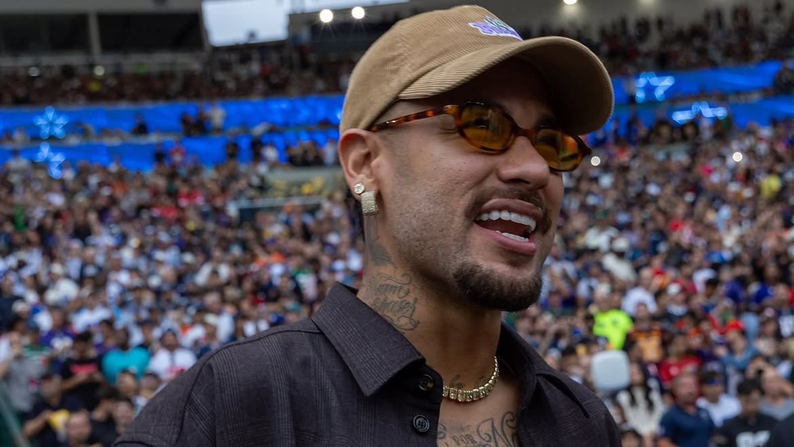 Close-up de Neymar sorrindo em estádio lotado, usando boné, óculos e corrente