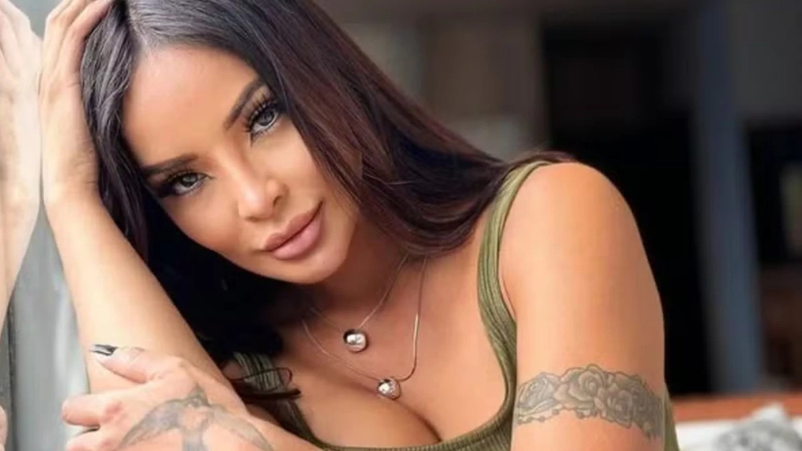 Close-up de Carol Nakamura sorrindo, cabelo escuro, camiseta verde e tatuagem floral no braço