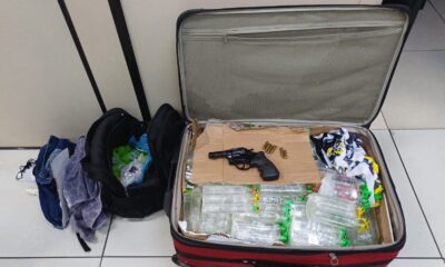 Mala aberta com revólver, munições, frascos transparentes; mochila e roupas no chão.