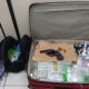 Mala aberta com revólver, munições, frascos transparentes; mochila e roupas no chão.