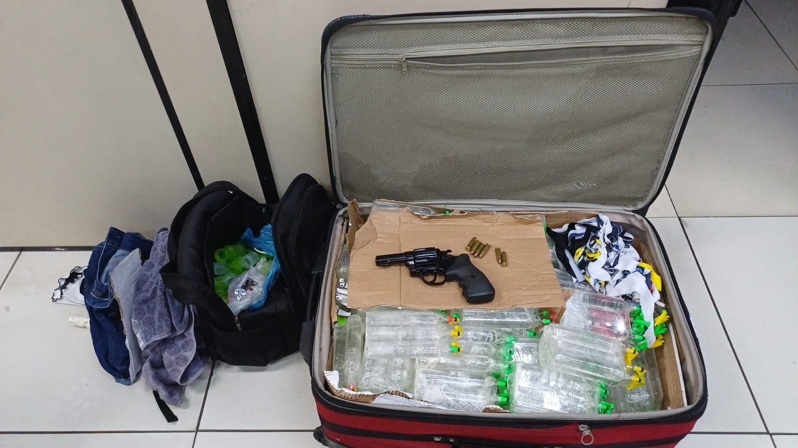 Mala aberta com revólver, munições, frascos transparentes; mochila e roupas no chão.