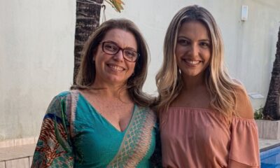Duas mulheres sorridentes: uma de óculos, blusa verde; a outra de cabelo loiro, blusa rosa