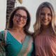 Duas mulheres sorridentes: uma de óculos, blusa verde; a outra de cabelo loiro, blusa rosa