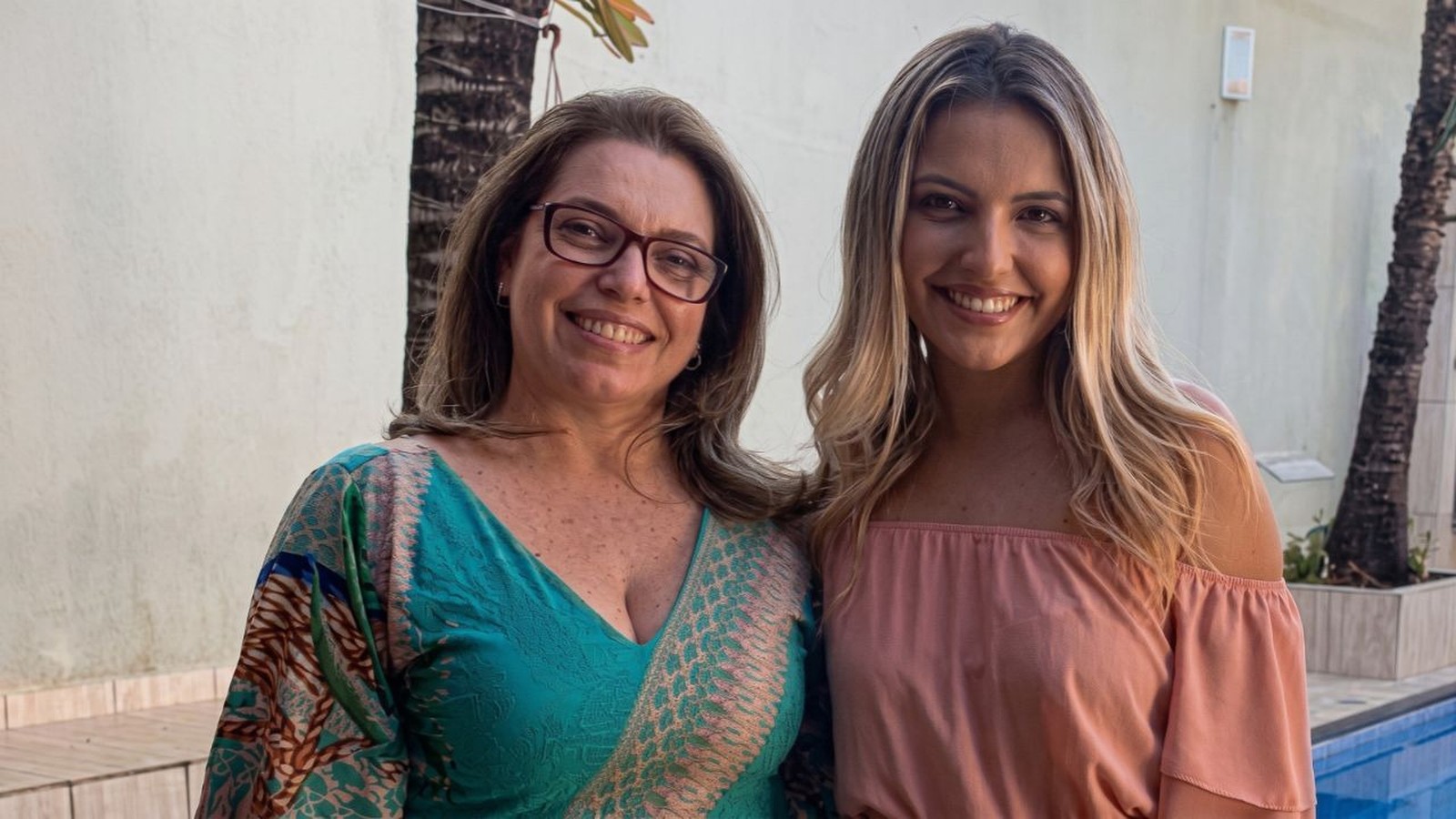 Duas mulheres sorridentes: uma de óculos, blusa verde; a outra de cabelo loiro, blusa rosa