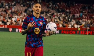 Raphinha em campo com camisa xadrez azul e vermelha, segurando uma bola de futebol