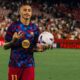 Raphinha em campo com camisa xadrez azul e vermelha, segurando uma bola de futebol