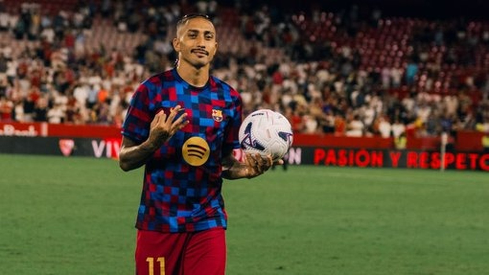 Raphinha em campo com camisa xadrez azul e vermelha, segurando uma bola de futebol
