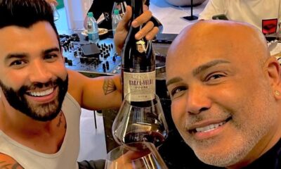 Dois homens sorridentes, um com barba e outro careca, brindam com vinho tinto e garrafa Barca-Velha 2004