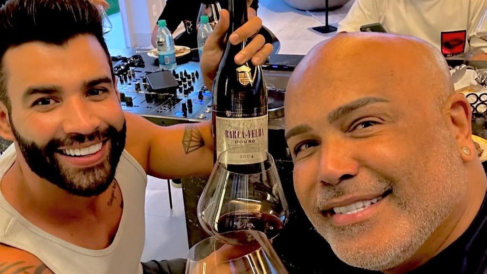 Dois homens sorridentes, um com barba e outro careca, brindam com vinho tinto e garrafa Barca-Velha 2004