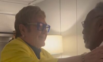 Elton John, óculos azuis e jaqueta amarela, cumprimenta Gilberto Gil em ambiente interno
