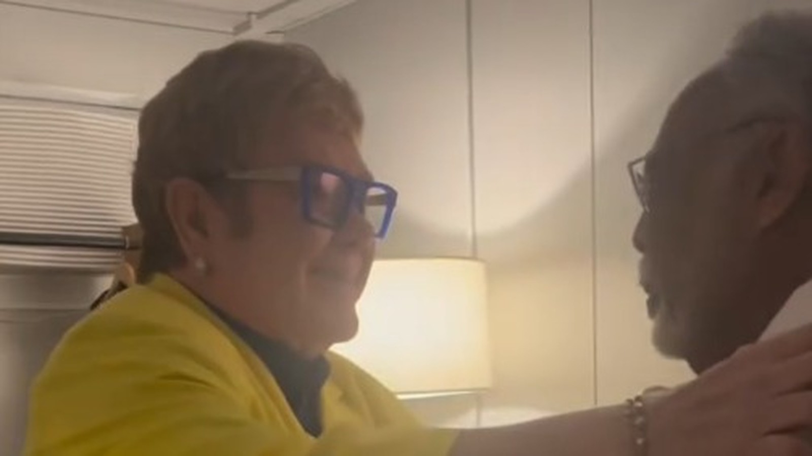 Elton John, óculos azuis e jaqueta amarela, cumprimenta Gilberto Gil em ambiente interno