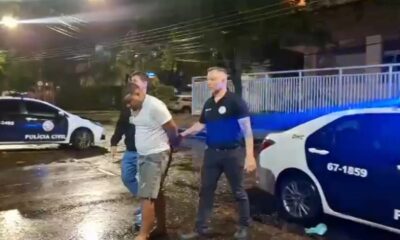 Homem algemado é conduzido por dois policiais civis ao lado de viaturas à noite