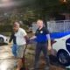 Homem algemado é conduzido por dois policiais civis ao lado de viaturas à noite