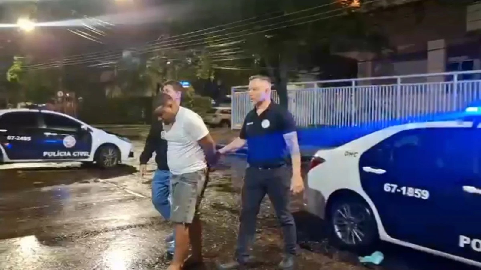 Homem algemado é conduzido por dois policiais civis ao lado de viaturas à noite