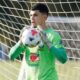 Jovem goleiro de camisa verde com logos Brasil e Guaraná, luvas, segura bola frente rede