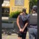Policial escolta homem algemado visto de costas em frente à 81ª Delegacia de Polícia