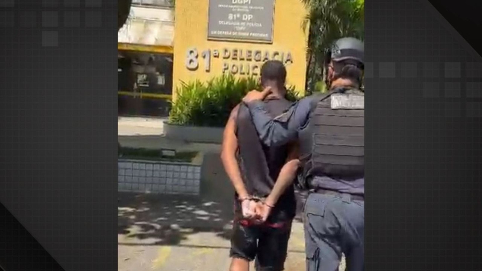 Policial escolta homem algemado visto de costas em frente à 81ª Delegacia de Polícia