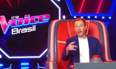 Tiago Leifert sentado na cadeira de técnico do The Voice Brasil, com botão vermelho e logo do programa