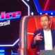 Tiago Leifert sentado na cadeira de técnico do The Voice Brasil, com botão vermelho e logo do programa