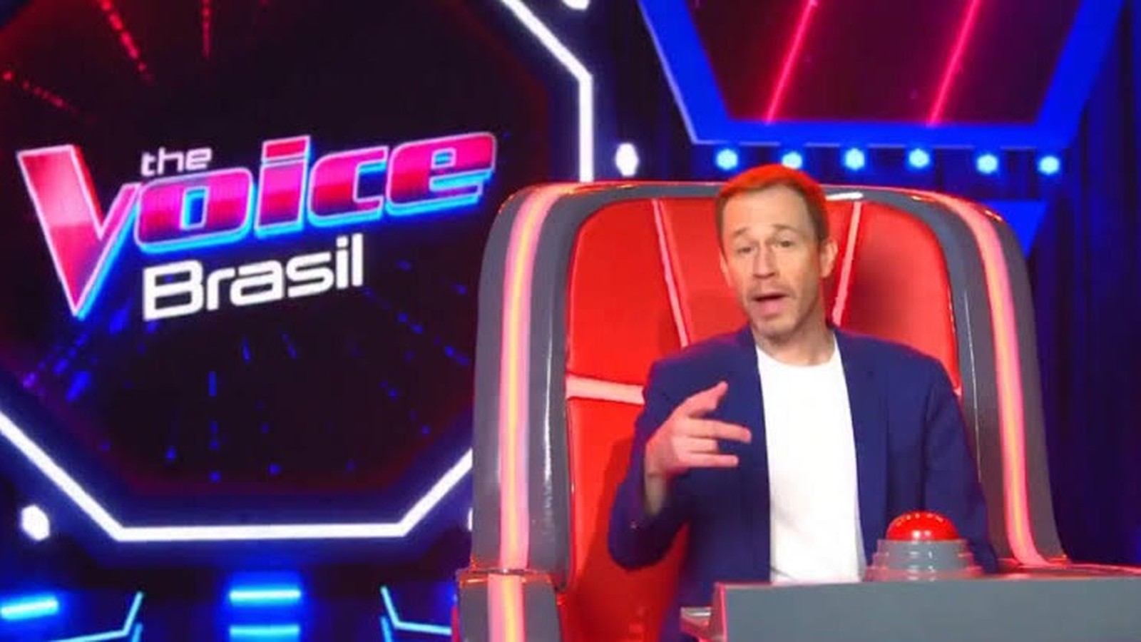 Tiago Leifert sentado na cadeira de técnico do The Voice Brasil, com botão vermelho e logo do programa