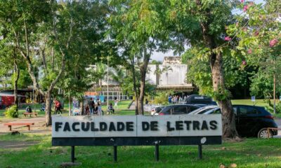 Sinal "FACULDADE DE LETRAS" em campus universitário com árvores, pessoas e carros