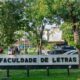 Sinal "FACULDADE DE LETRAS" em campus universitário com árvores, pessoas e carros