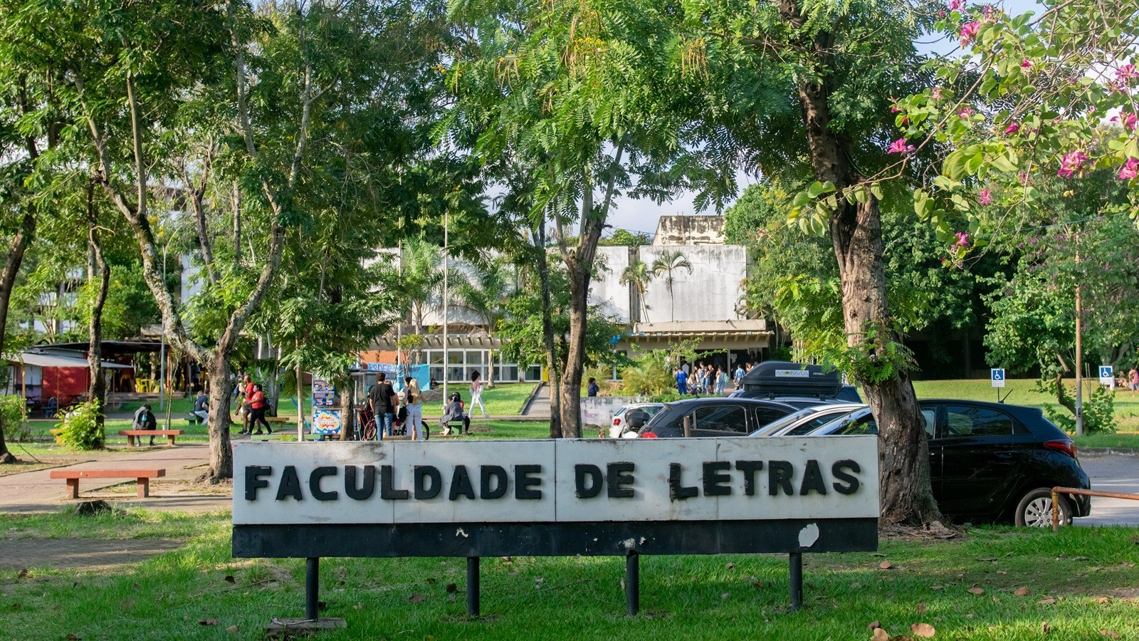 Sinal "FACULDADE DE LETRAS" em campus universitário com árvores, pessoas e carros
