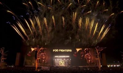 Palco iluminado do Foo Fighters com logo e pirotecnia dourada no céu noturno, com plateia abaixo