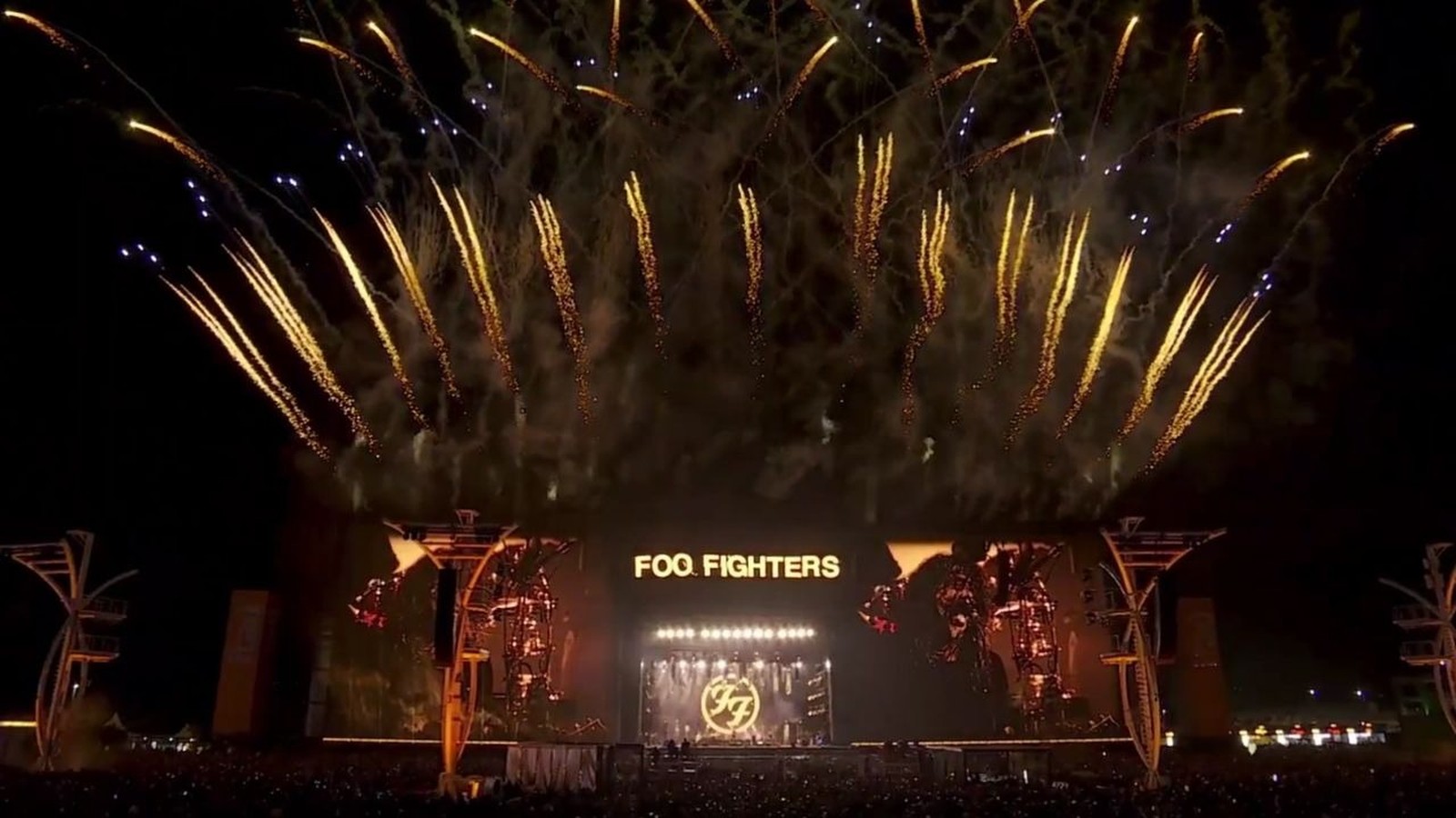 Palco iluminado do Foo Fighters com logo e pirotecnia dourada no céu noturno, com plateia abaixo