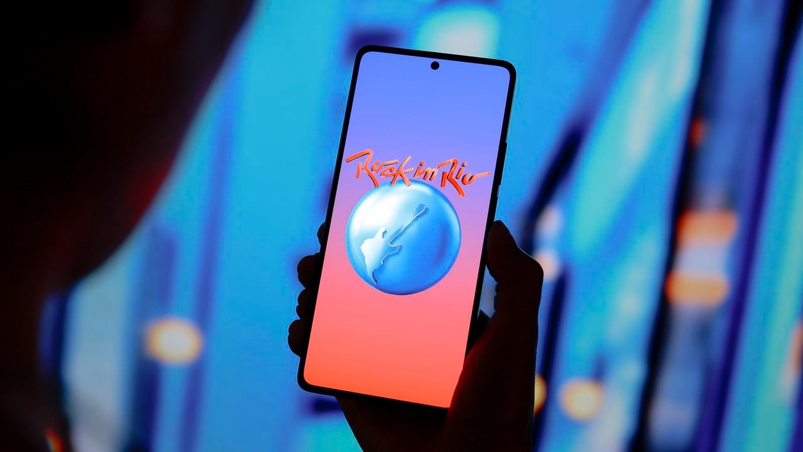 Mão segura smartphone com logo 'Rock in Rio' e ilustração de globo com guitarra na tela.