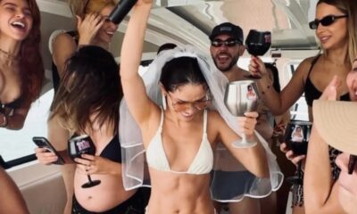 Mulher com véu de noiva e biquíni branco, braços erguidos, celebrando em barco com amigos