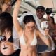 Mulher com véu de noiva e biquíni branco, braços erguidos, celebrando em barco com amigos