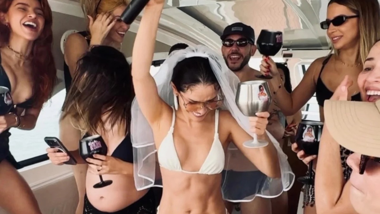 Mulher com véu de noiva e biquíni branco, braços erguidos, celebrando em barco com amigos