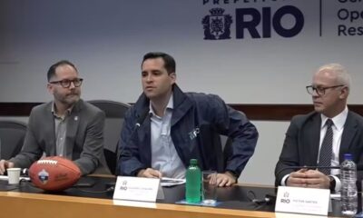 Três homens em coletiva de imprensa, com Victor Santos à direita e uma bola de futebol americano na mesa