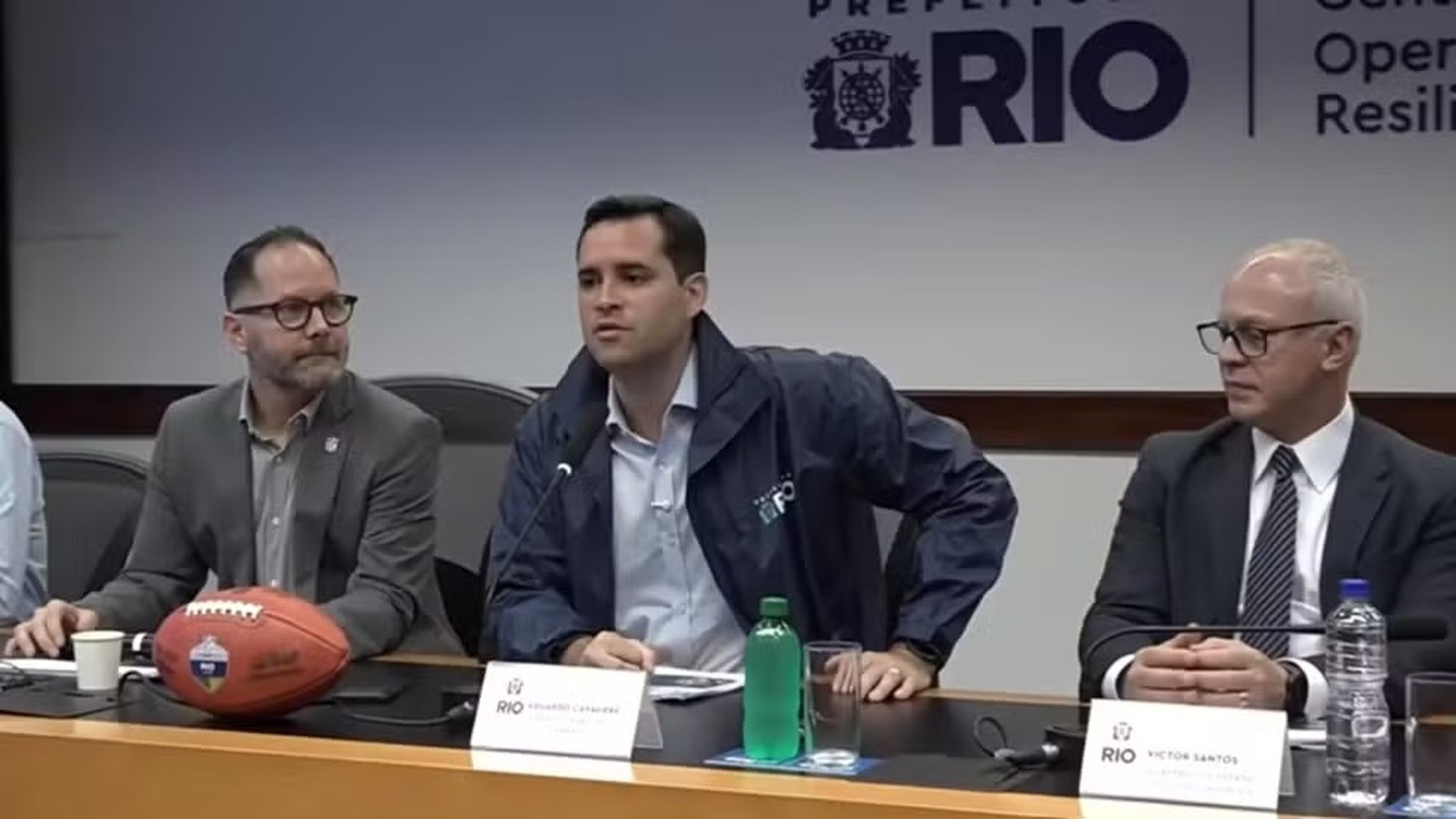 Três homens em coletiva de imprensa, com Victor Santos à direita e uma bola de futebol americano na mesa