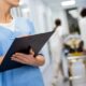 Mulher em uniforme médico azul escrevendo em prancheta no corredor de um hospital