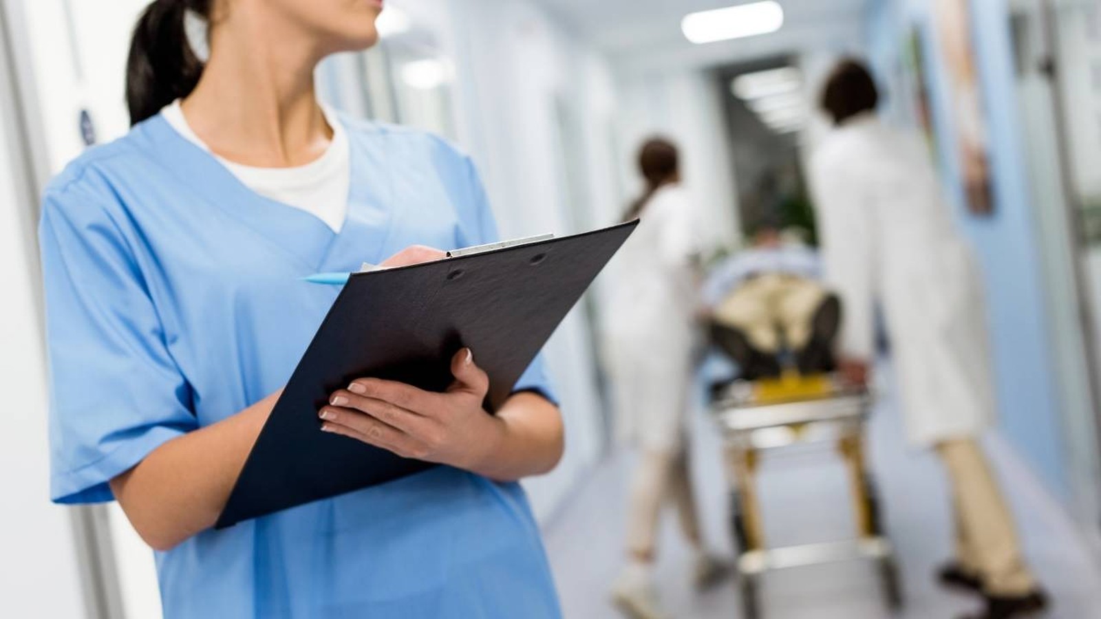 Mulher em uniforme médico azul escrevendo em prancheta no corredor de um hospital