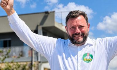 Candidato Dr. Elcio Kuhnen sorrindo, braços erguidos, camisa branca, broche '15777'