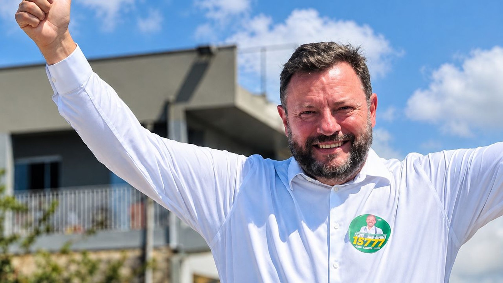 Candidato Dr. Elcio Kuhnen sorrindo, braços erguidos, camisa branca, broche '15777'