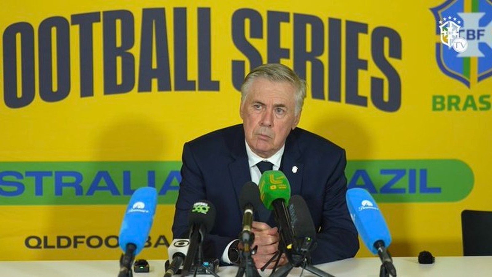 Carlo Ancelotti, terno azul, microfones, fundo amarelo com "FOOTBALL SERIES AUSTRALIA BRAZIL CBF TV"