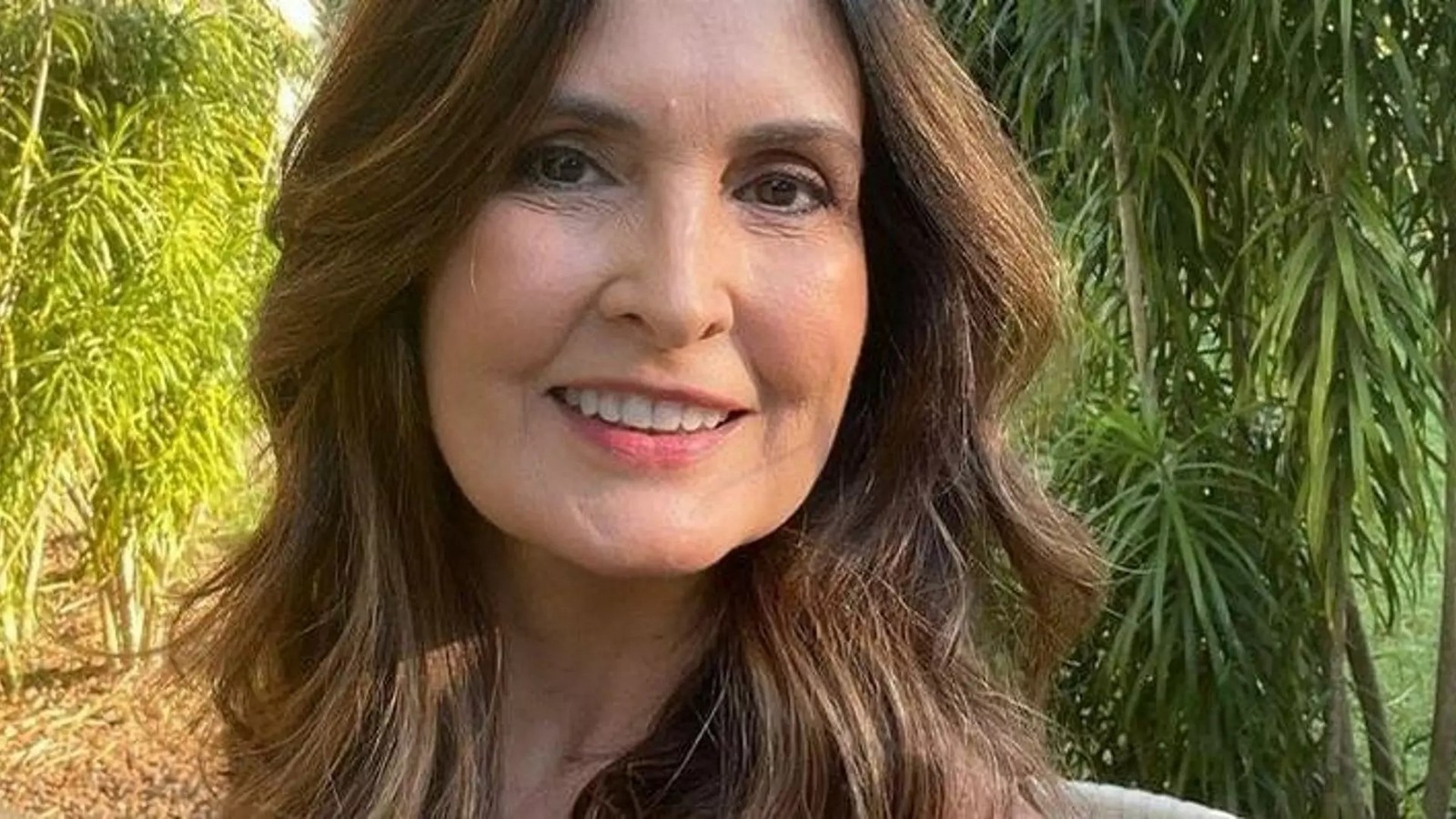 Fátima Bernardes sorrindo em close-up, com cabelos castanhos e fundo de folhagem verde