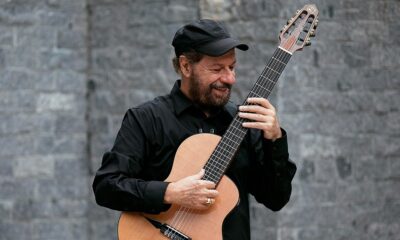 Homem sorridente de barba, chapéu e camisa pretos toca violão clássico