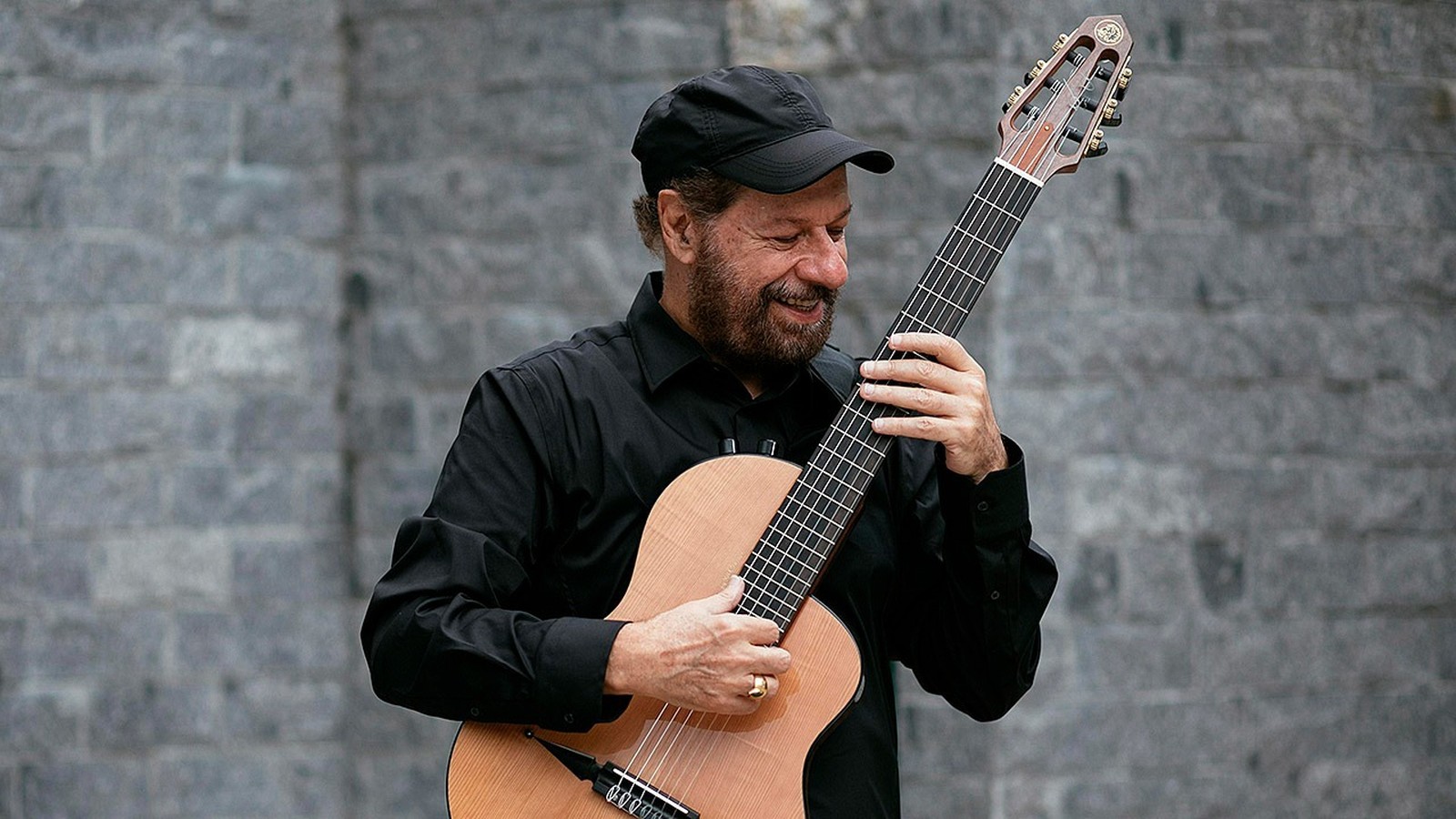 Homem sorridente de barba, chapéu e camisa pretos toca violão clássico