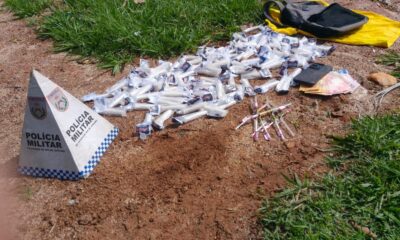 Pirâmide da Polícia Militar, drogas, seringas, dinheiro, celular e mochila sobre o chão de terra