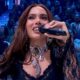 Anitta sorrindo, segurando microfone, com colar e blusa preta de renda, plateia ao fundo