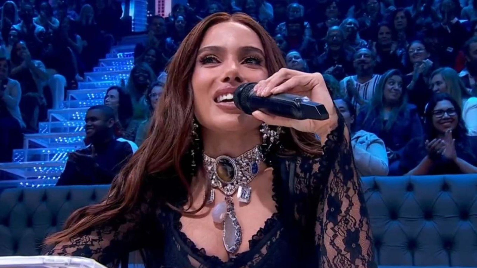 Anitta sorrindo, segurando microfone, com colar e blusa preta de renda, plateia ao fundo