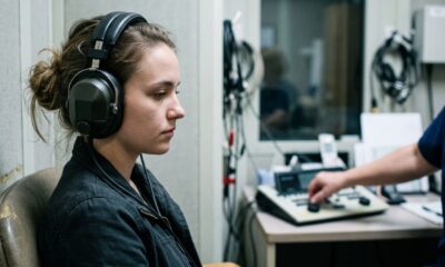 Mulher jovem com fones de ouvido pretos faz exame de audiometria em cabine à prova de som