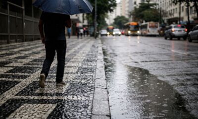 Homem com guarda-chuva caminha em calçada molhada, rua com poças e carros desfocados