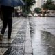 Homem com guarda-chuva caminha em calçada molhada, rua com poças e carros desfocados