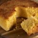 Bolo de fubá cremoso e dourado com uma fatia removida, sobre uma tábua de madeira